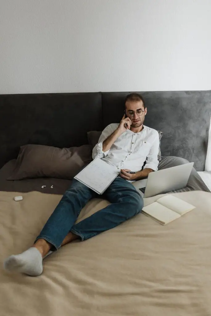 a man in white long sleeves and denim jeans resting on the bed while having a phone call