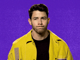 Video gif. Singer Nick Jonas, wearing a bright yellow jacket, sheepishly raises his hand and looks around.